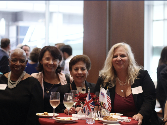Six women stand smiling behind a table topped with food, drinks, and various nation's flags