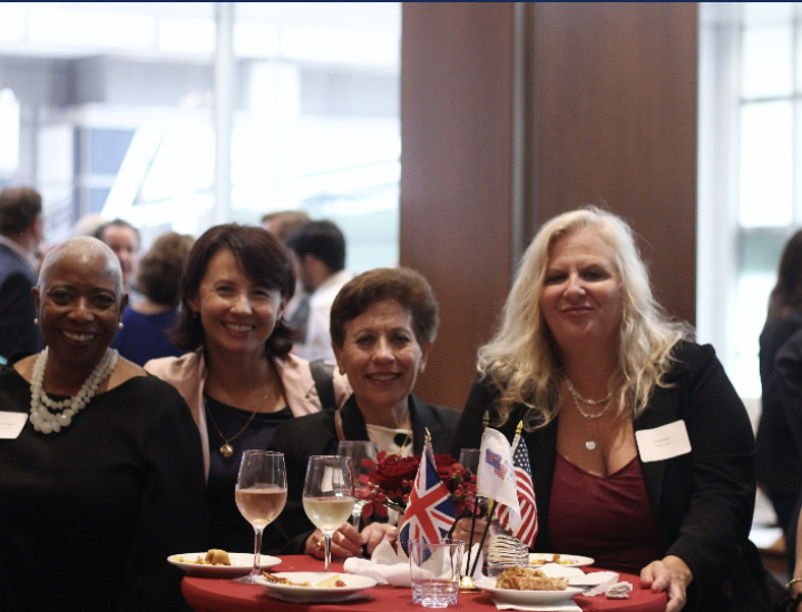 Six women stand smiling behind a table topped with food, drinks, and various nation's flags